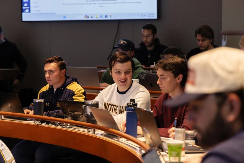 Students sit in curved rows within a tiered, modern lecture hall, working on laptops during class. In the center, a male student in a white Notre Dame crewneck sweatshirt smiles broadly while typing. To his left, a classmate wears a navy-and-gold jacket, while to his right, another student in a maroon sweatshirt takes notes. A large projection screen displays a web page on the wall above them.