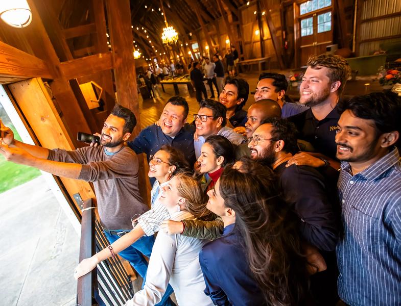 A diverse group of young adults huddles closely together inside a spacious event venue featuring high, vaulted wooden beams and warm chandeliers. On the left, a man holds up a smartphone to snap a group selfie while everyone leans in, smiling and laughing toward the camera. The attendees are dressed in smart-casual attire, enjoying a lively social gathering against the background of exposed timber architecture.