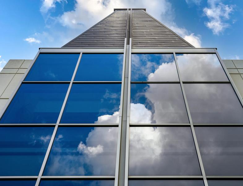 A symmetrical, low-angle photograph looking straight up at the façade of a modern architectural structure. Large, grid-patterned glass windows dominate the lower half, clearly reflecting bright white clouds and a blue sky above. A slatted, metallic central spire rises sharply between the window sections, tapering upward against a sky filled with scattered clouds.