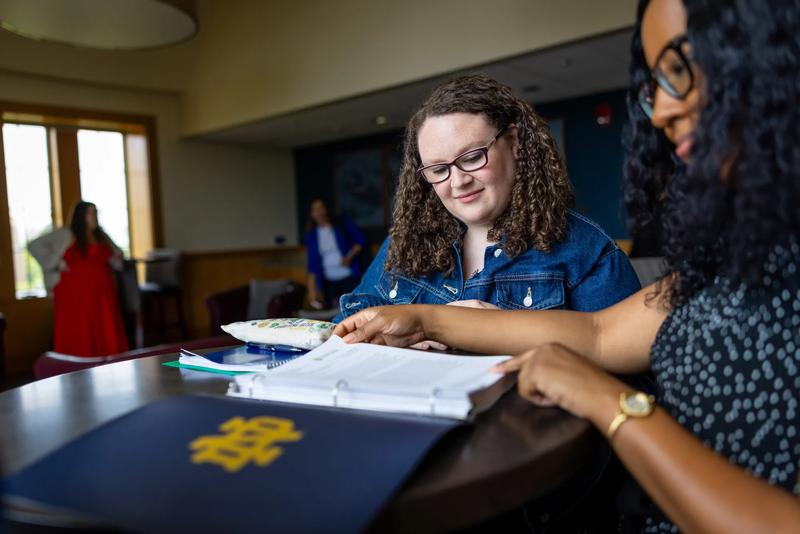 A close-up view of two women collaborating over study materials at a dark wood table. In the foreground on the right, a woman with dark curly hair and glasses points to a open three-ring binder next to a navy blue folder stamped with a gold Notre Dame "ND" monogram. Across from her, a woman with curly brown hair, glasses, and a denim jacket looks down intently at the document.