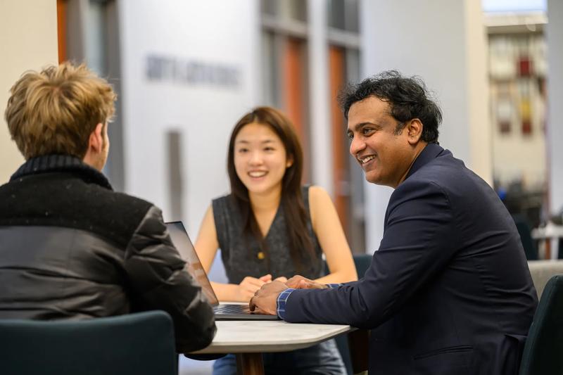 Three people are gathered around a table speaking together. The environment is a college meeting space with a standing table. Doors and hall space can be seen in the background.