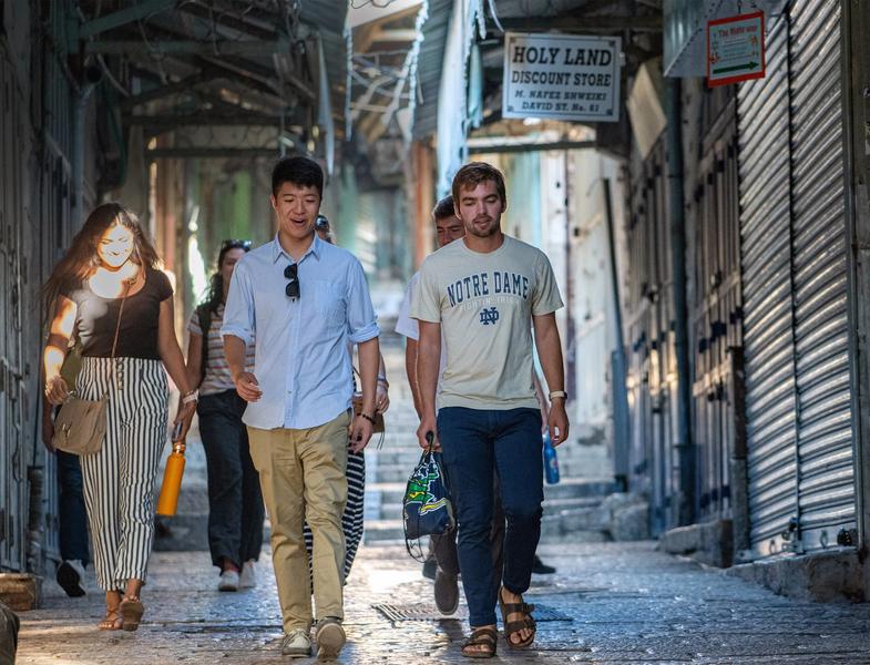 A group of young adults walk toward the camera through a narrow, cobblestone alley in an old city market. In the center, a man wearing a light blue button-down shirt and khaki pants walks alongside a man in an off-white "Notre Dame" t-shirt, blue jeans, and sandals. To the left, a woman in a black top and black-and-white striped pants holds a bright yellow water bottle. In the background, arched stone structures overhang the street, and a overhead sign reads "HOLY LAND DISCOUNT STORE." Closed metal corrugated shop shutters line the right side of the path.