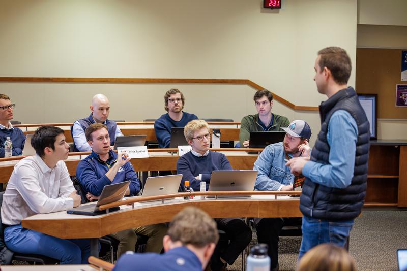 An instructor wearing a dark quilted vest over a blue shirt stands at the front of a tiered, amphitheater-style classroom, addressing a group of students. The students sit at curved wooden desks equipped with laptops and name cards, with one male student in a navy pullover actively speaking while raising his hand to make a point. The surrounding classmates listen attentively as the class discussion takes place against neutral beige walls.