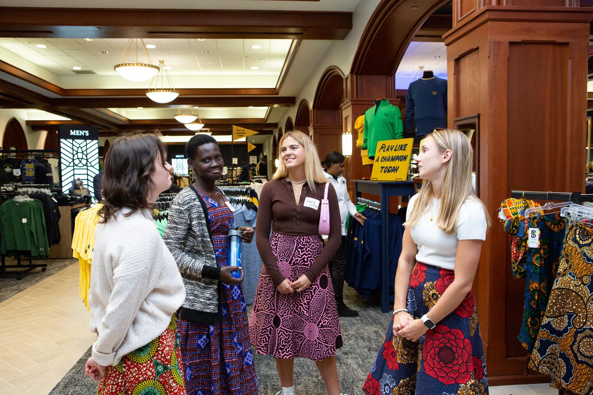 Four young women stand in a circle in the Hammes bookstore, facing each other and talking. They are all wearing Ugandan skirts with brightly-colored patterns, and more Ugandan skirts hang on the display rack to the right. Behind them, the Notre Dame bookstore features wood paneling and racks of navy, gold, and green apparel. | Photo by Jennifer Mayo/University of Notre Dame