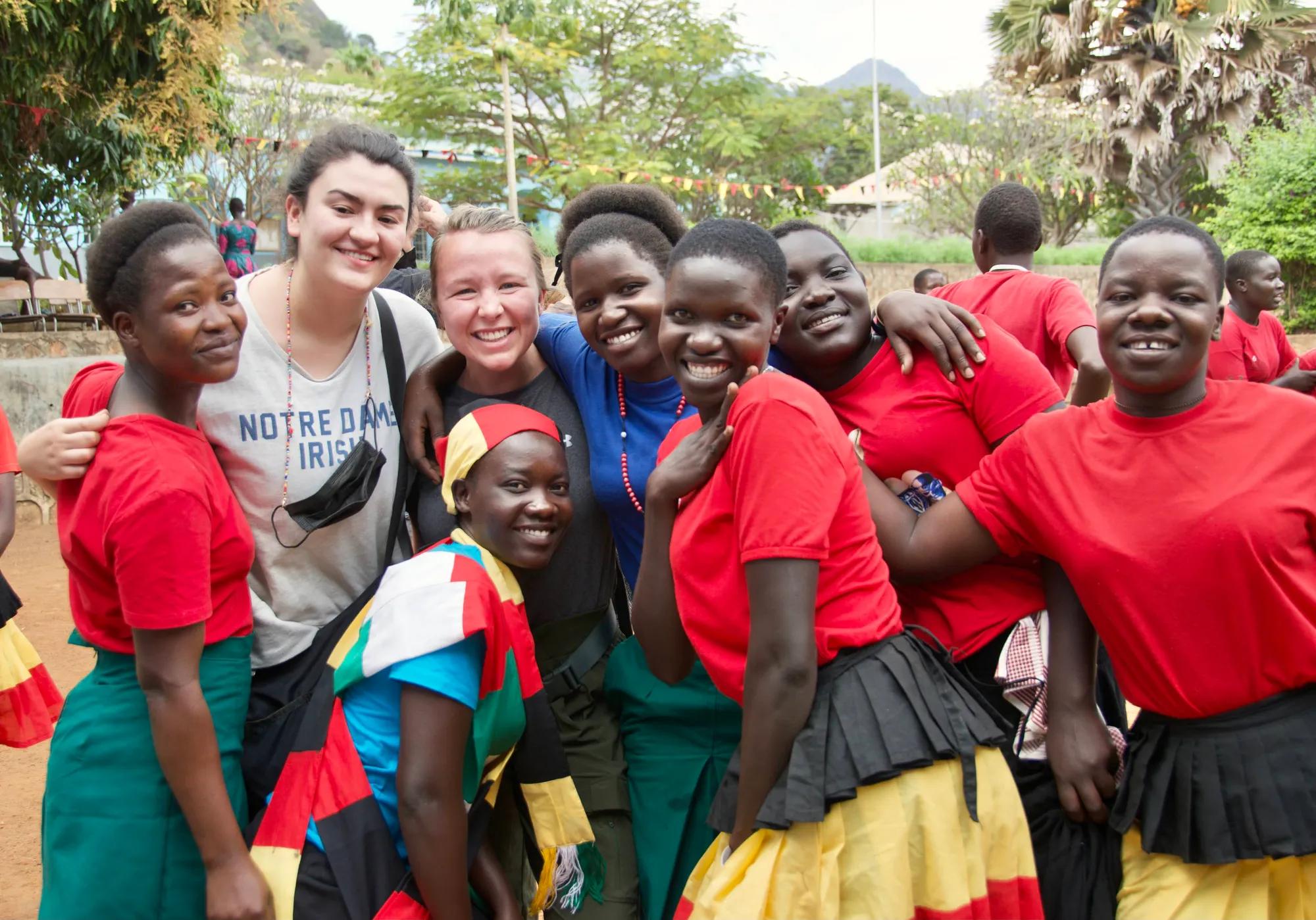 Eight young women pose for a photo, hugging and smiling. Two women are Notre Dame students and wear casual t-shirts and clothing, while the other six women are Ugandan and wear traditional skirts and attire in red, yellow and black colors, with matching red t-shirts. In the background, leafy green trees fill the frame, while the outline of thatched roofs and small mountains can be seen through the trees. 