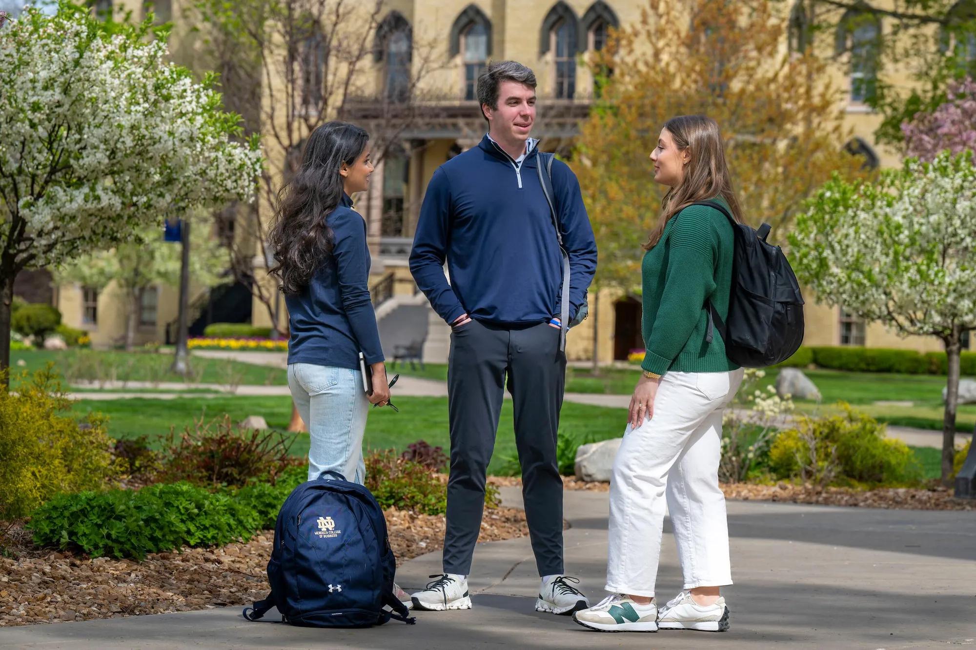 Three students discussing on the sidewalk in front of a building, capturing a moment of interaction in an educational setting.