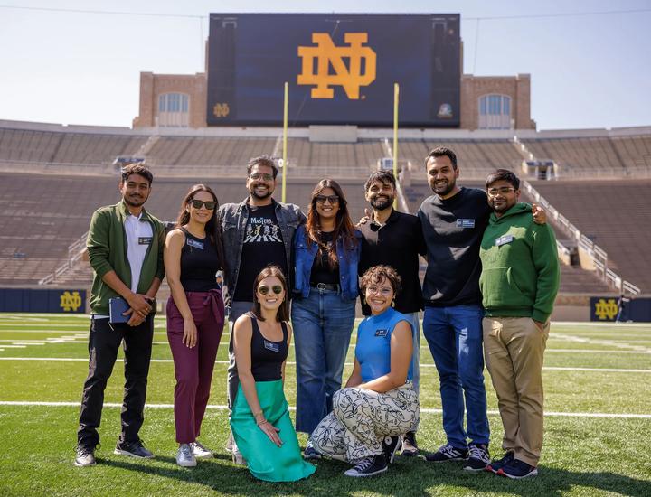 Eight students pose for a group photo on the green turf of Notre Dame Stadium on a bright, sunny day. The massive stadium jumbotron in the background proudly displays a large, gold interlocking Notre Dame "ND" monogram. The students, each wearing official Notre Dame name badges, are dressed in smart casual attire and arranged in two rows, smiling together on the field.