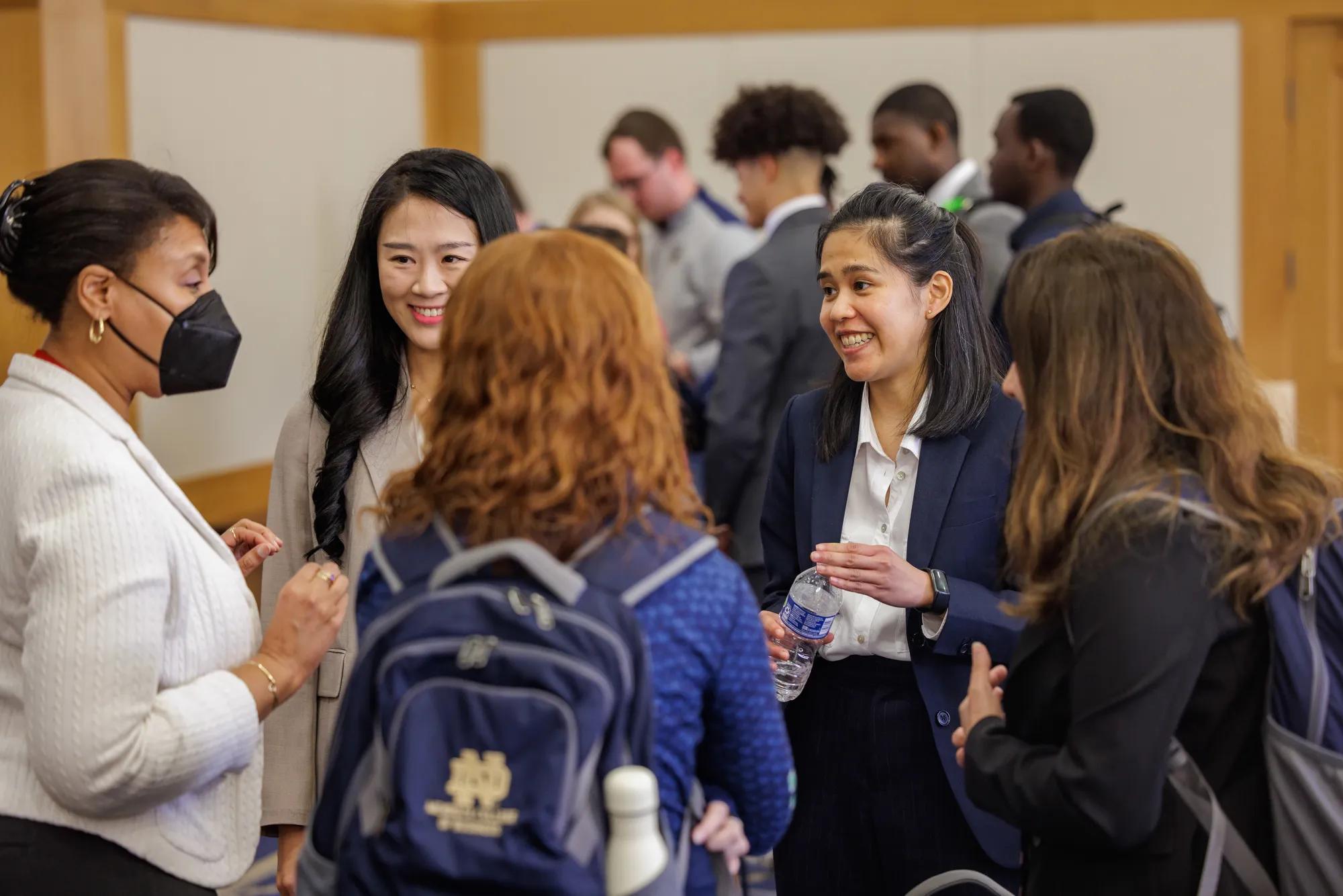 Four female students stand and talk with a female Mendoza staff member as part of a lunch and networking social. The two students facing the camera are smiling and focused on the staff member as she talks. In the background, other students can be seen standing in circles having similar networking conversations. | Photo by Peter Ringenberg/University of Notre Dame