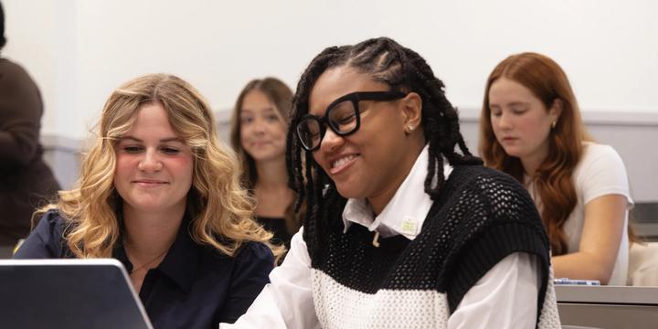 Two female students smile as they look at a laptop screen together while sitting at a desk in a classroom setting. The student on the right, wearing black-framed glasses, locs, and a black-and-white knit vest over a white collared shirt, operates the laptop. Beside her, a blonde student in a dark top smiles approvingly. Other students sit in the background of the bright classroom.