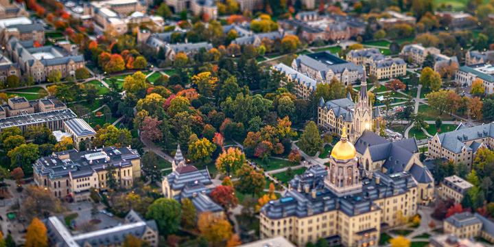 An elevated, high-angle tilt-shift photograph showing a university campus framed by autumn colors. The selective focus centers on the Main Building with its bright Golden Dome and the neighboring spire of the Basilica of the Sacred Heart. Surrounding the historic stone buildings are quads, walkways, and dense trees displaying rich yellow, orange, red, and green foliage, with the top and bottom edges softly blurred.
