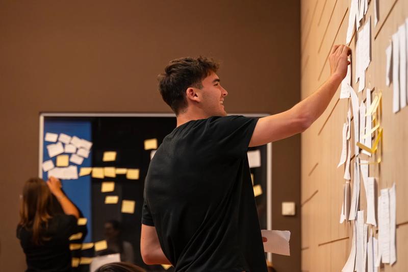 A young man in a black t-shirt smiles as he places a note on a wall covered with papers, while another person does the same in the background.