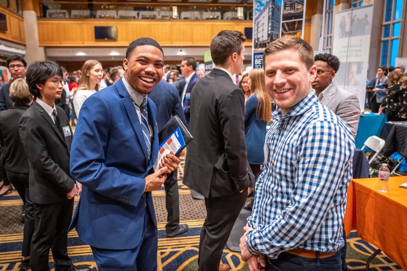 A vibrant career fair scene with a diverse group of attendees networking in a professional setting. In the foreground, a young man in a blue suit smiles while holding a portfolio and a brochure, engaging with a recruiter in a checkered shirt. The background features other students and professionals conversing, with company booths and banners visible throughout the event.