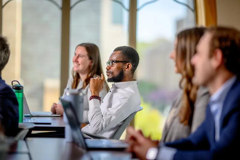 ND MBA students sitting at a conference table, attentively listening. A man in a white shirt is in focus, with others blurred in the foreground.