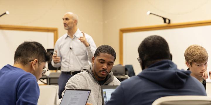 In a classroom setting, four students sit around a table focusing on laptops and taking notes during a lecture. In the foreground, a student in a grey zip-up hoodie writes in a notebook beside an open laptop. In the soft-focus background, an instructor in a light button-down shirt stands in front of whiteboards, gesturing with his hand as he presents to the class.