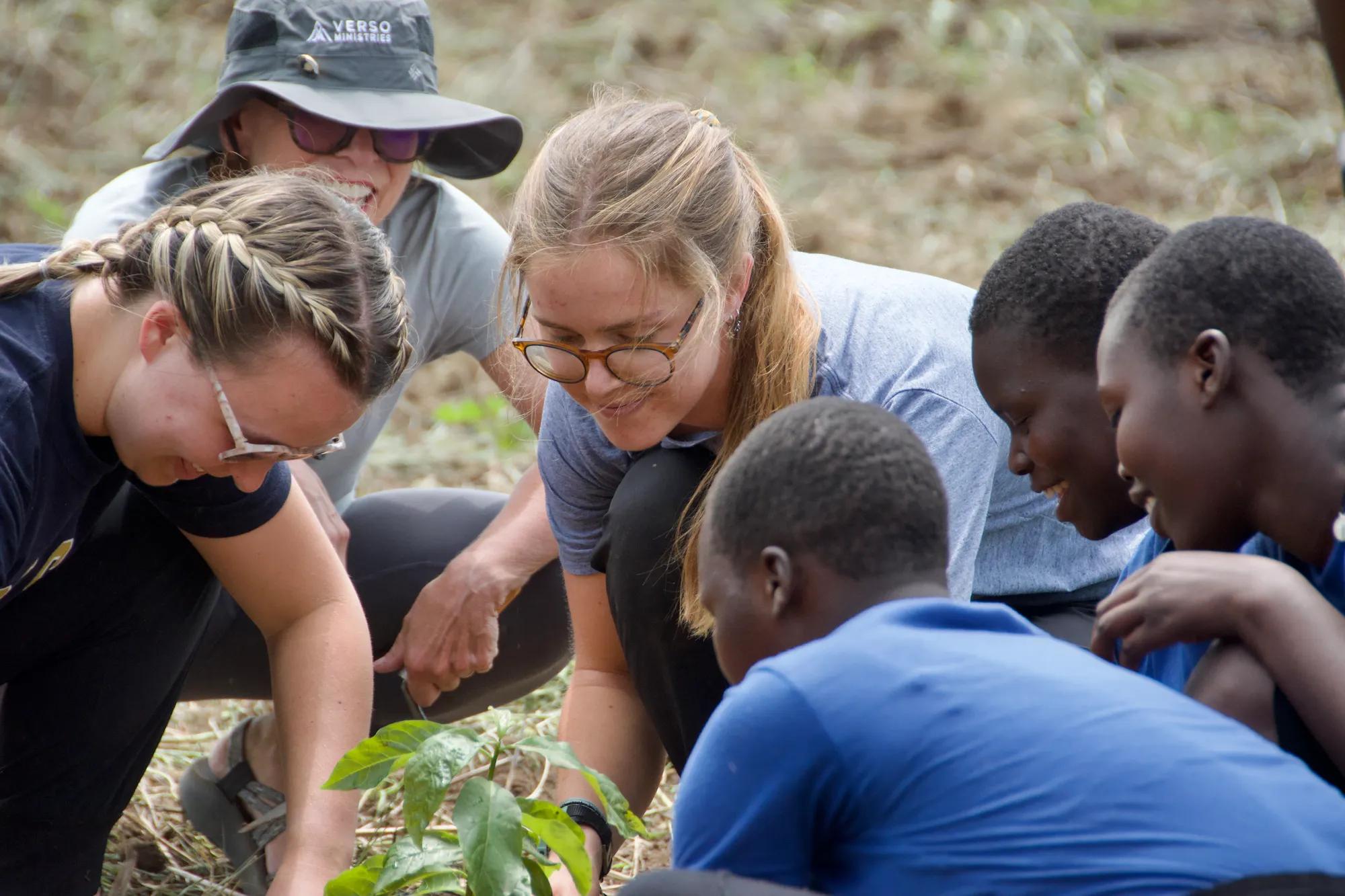 Three female Notre Dame students, all with glasses and casual t-shirts, crouch alongside three young women from Saint Bakhita Vocational Training Center to plant a tree. They are all smiling and reaching towards the plant, helping pack dirt in around the sapling. A light brown straw-covered field fills the background. 