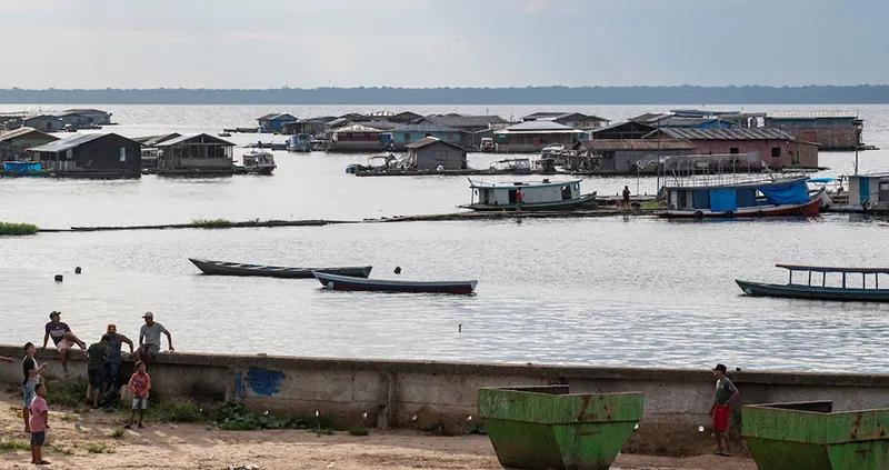 Floating houses on the Amazon river in Brazil with boats nearby. People stand on a concrete embankment in the foreground under a cloudy sky.