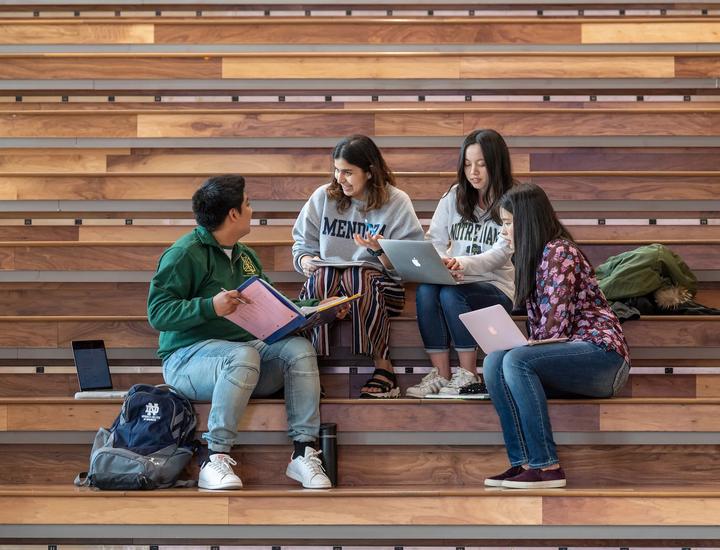 our students sit together on broad, indoor wooden tiered steps while studying and conversing. On the left, a student wearing a green jacket holds an open binder next to a navy blue Notre Dame backpack. Beside him, a student in a grey "MENDOZA" sweatshirt gestures while speaking. Next to her, a student in a white "NOTRE DAME" sweatshirt works on a laptop, alongside a fourth student on the right typing on a second laptop.