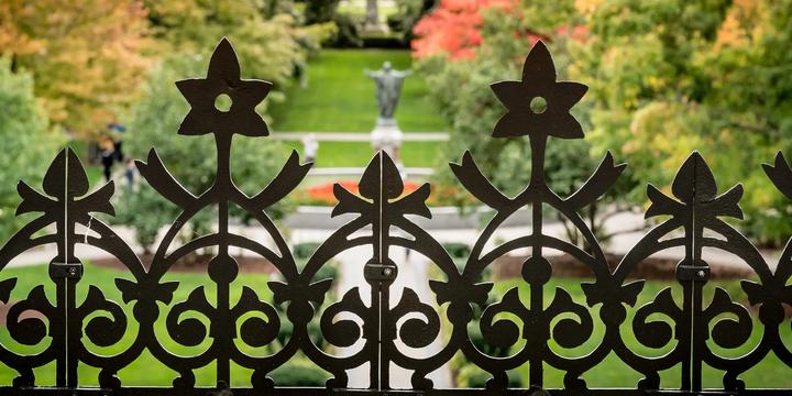 A close-up shot of a decorative dark wrought-iron fence featuring intricate scrollwork and star-like finials. In the softly blurred background, a green lawn stretches down a campus quad bordered by trees exhibiting vibrant fall foliage in red, orange, and green, leading toward a central statue on a pedestal.