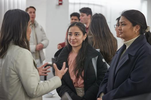A group of attendees and students engage in small group conversations during an indoor gathering or site visit. In the foreground, a woman in a light jacket gestures with her hands as she speaks to two women—one wearing a black puffer coat over a pink top and the other in a navy overcoat—who listen attentively. Additional attendees converse in the softly blurred background.