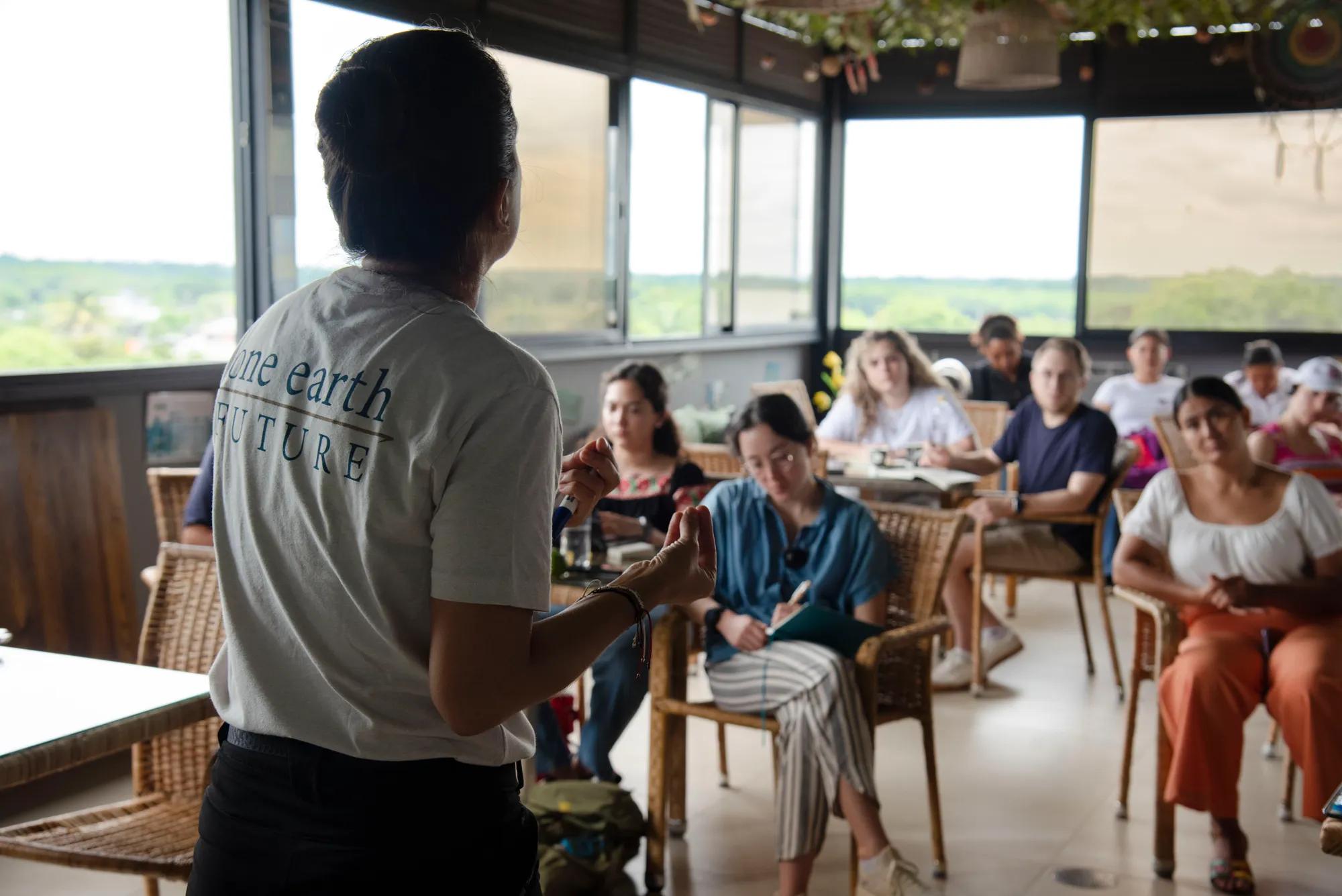 nine students seated in wicker chairs listen intently to a speaker at the front of a large open room lined with windows peering out over a field