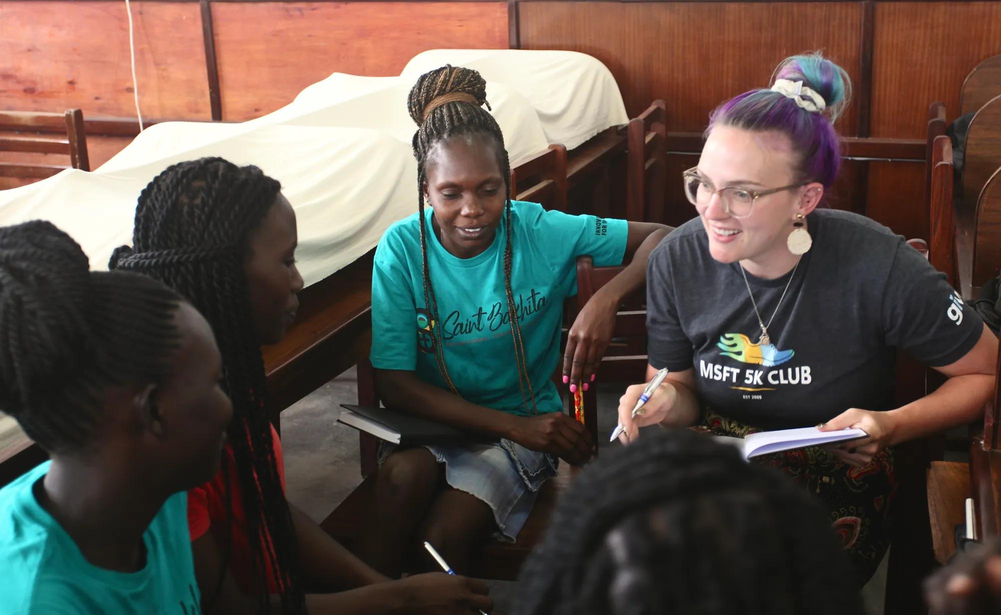 Five young women, one from Notre Dame and the other four from St. Bakhita Vocational Training Center, sit in a wood-paneled room on wooden chairs in a circle. The Notre Dame student holds a pen and notebook and is smiling at one of the SBVTC students as they are actively engaged in a group discussion.