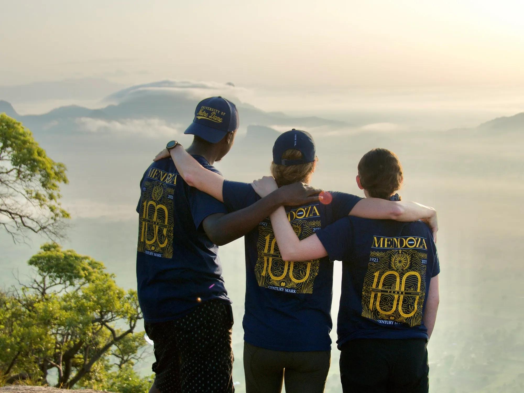 One young man and two young women stand with their backs to the camera and their arms around each other's shoulders. They are dressed in Mendoza 100th anniversary t-shirts and gaze out over a bright and landscape with leafy green trees on the left and small mountains in front, with clouds rolling over the landscape. 