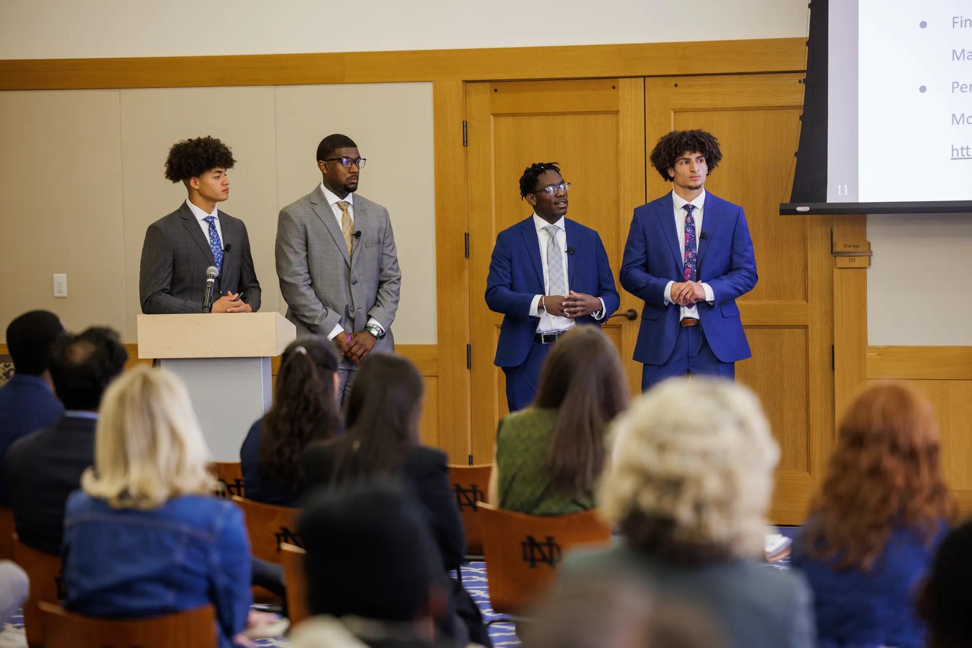 A team of four male students in business professional attire stand in front of the audience and judges to present their case solution. | Photo by Peter Ringenberg/University of Notre Dame