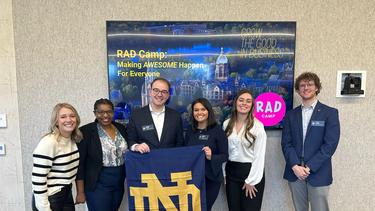 Six professionals stand side-by-side indoors in front of a large digital display wall showing campus imagery alongside the text "RAD Camp: Making AWESOME Happen For Everyone" and the Mendoza motto "GROW THE GOOD IN BUSINESS." In the center, two team members hold up a navy blue Notre Dame "ND" flag with gold lettering and a shamrock icon in the corner. The group is dressed in professional business attire, including suits, blazers, and dark trousers.
